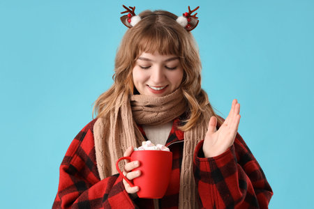 Pretty young woman in scarf holding cup of hot chocolate with marshmallows on blue backgroundの写真素材
