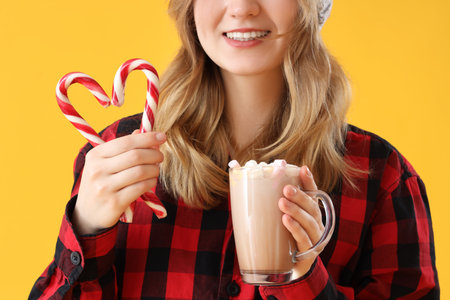 Young woman holding cup of hot chocolate with marshmallows and candy canes on yellow background, closeupの写真素材
