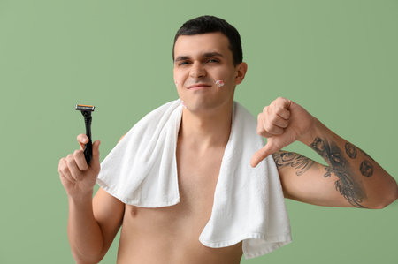 Handsome displeased man with towel, razor and cuts on skin after shaving showing thumb-down gesture against green backgroundの写真素材
