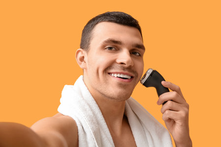 Handsome young man with towel and electric shaver on yellow backgroundの写真素材