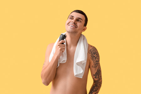 Handsome young man with towel and electric shaver on yellow backgroundの写真素材