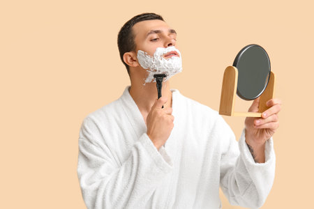 Handsome young man with mirror and razor shaving his face on beige backgroundの写真素材