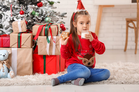 Cute little girl in Santa hat headband with sweet cookie and glass of milk at home on Christmas Eveの写真素材