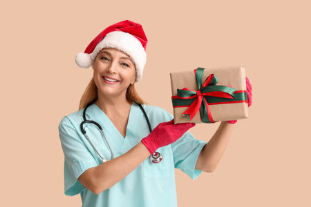 Mature female doctor in Santa hat with Christmas gift box on beige backgroundの写真素材