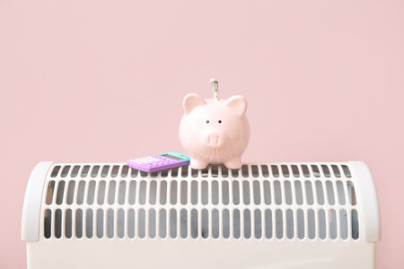 Piggy bank with calculator on electric convector heater near pink wall at home. Heating saving conceptの写真素材