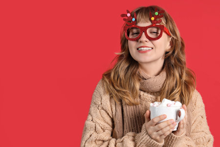 Pretty young woman in Christmas glasses holding cup of hot chocolate on red backgroundの写真素材