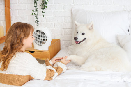 Young woman with Samoyed dog warming near electric fan heater in bedroomの写真素材