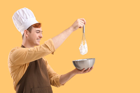 Male confectioner in apron with bowl and whiskey preparing tasty meringue on yellow backgroundの写真素材