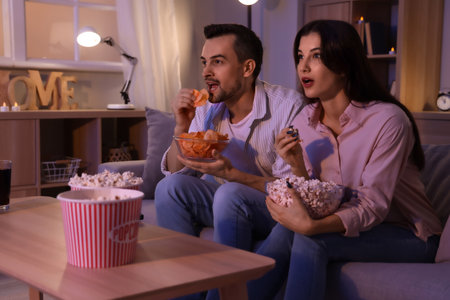 Young couple with potato chips and popcorn watching TV at home in eveningの写真素材