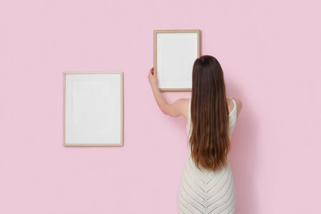 Young woman hanging blank frame on pink wall, back view