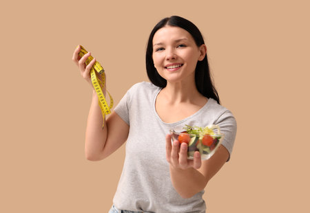 Beautiful young woman with measuring tape and bowl of fresh vegetable salad on beige background. Weight loss conceptの写真素材