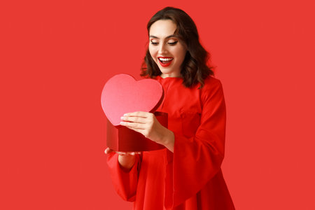 Surprised young woman with heart shaped gift box on red backgroundの写真素材