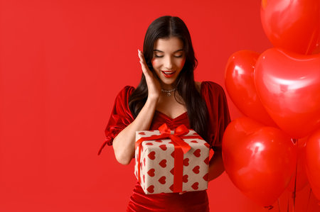 Surprised young woman with kiss marks on her face holding gift box and heart shaped air balloons on red backgroundの写真素材