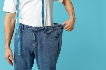 Handsome sporty young man in loose jeans with measuring tape on blue background, closeup. Weight loss conceptの写真素材