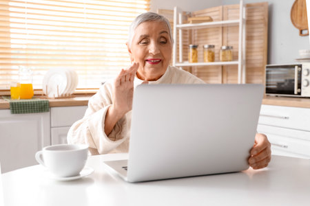 Senior woman with laptop video chatting at table in kitchenの写真素材