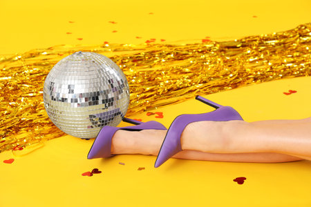 Female legs in stylish high heels with disco ball and confetti on yellow backgroundの写真素材