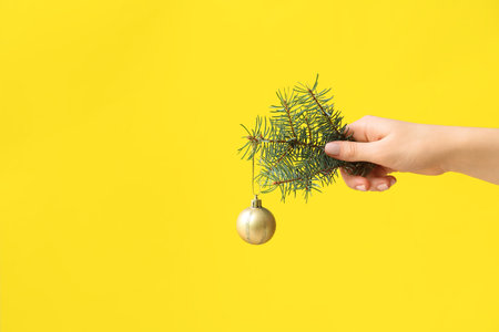 Female hand with fir branch and Christmas ball on yellow backgroundの写真素材