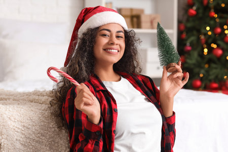 Beautiful young happy African-American woman with Santa hat, decorative tree and candy cane in bedroom decorated for Christmasの写真素材