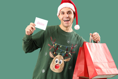 Handsome young happy man in Santa hat with shopping bags and gift card on green backgroundの写真素材