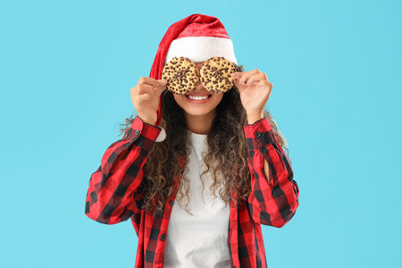 Beautiful young happy African-American woman in checkered pajamas and Santa hat with cookies on blue backgroundの写真素材