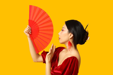 Beautiful young Asian woman with fan on yellow background. Chinese New Year celebrationの写真素材
