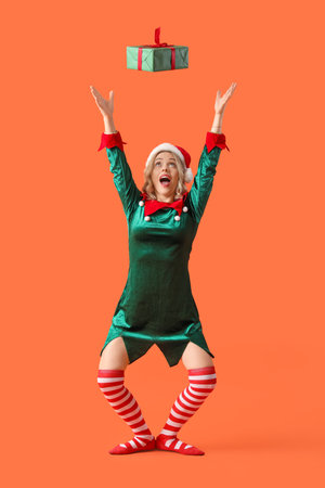 Happy young woman dressed as elf with Christmas gift on orange backgroundの写真素材