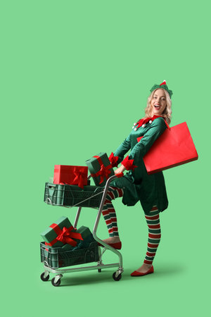 Happy young woman dressed as elf with shopping cart and Christmas gifts on green backgroundの写真素材