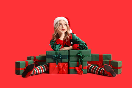Thoughtful young woman dressed as elf with Christmas gifts on red background