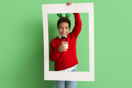 Little African-American boy in reindeer horns with glass cup of milk and frame on green backgroundの写真素材