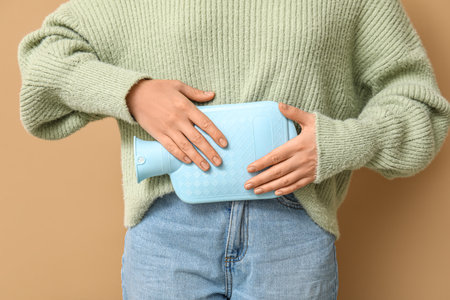 Young woman with hot water bottle having menstrual cramps on beige background, closeupの写真素材