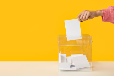Hand putting paper voting in ballot box on yellow backgroundの写真素材