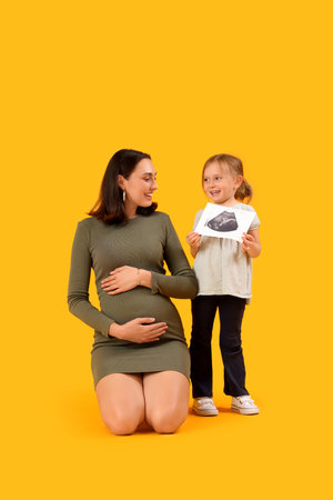 Beautiful pregnant mother and her cute little daughter with sonogram image sitting on yellow backgroundの写真素材