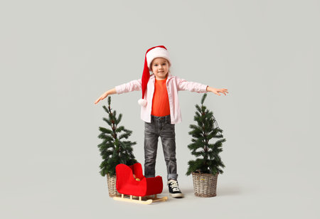 Cute little girl in Santa hat with Christmas trees and sledge on light backgroundの写真素材