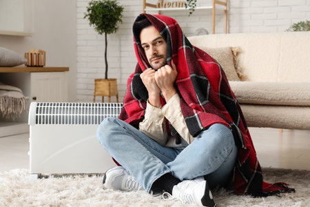 Sick young man with plaid warming near radiator at home