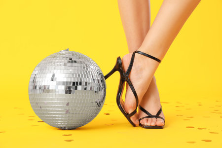Female legs in stylish high heels with disco ball and confetti on yellow backgroundの写真素材