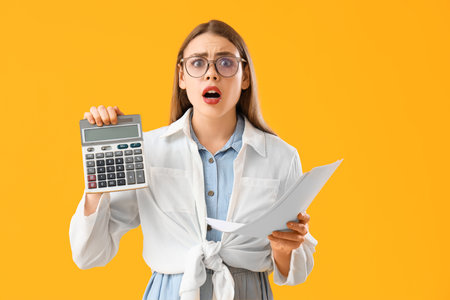 Shocked young woman with calculator and bills on yellow background. Price rise conceptの写真素材
