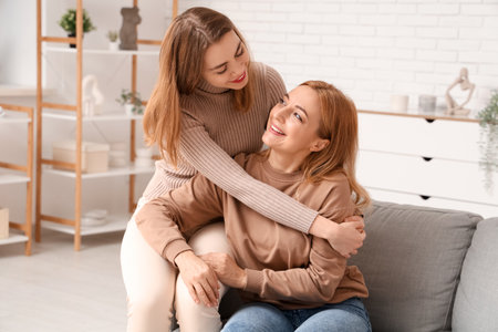 Beautiful young woman hugging her mother on sofa at home. International Hug Dayの写真素材