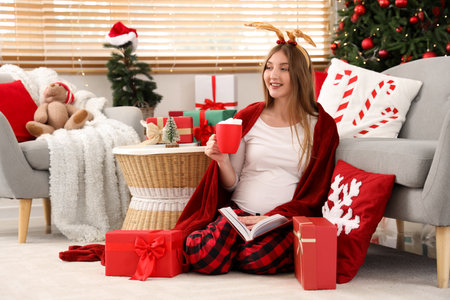 Young pregnant woman with deer horns, gift boxes and cup of cocoa reading book in room decorated for Christmasの写真素材
