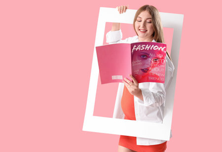 Young happy pregnant woman with magazine and frame on pink backgroundの写真素材