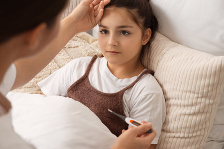 Sick little girl and her mother with thermometer in bedroomの写真素材
