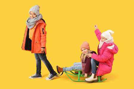 Cute children in winter clothes with sledge on yellow backgroundの写真素材