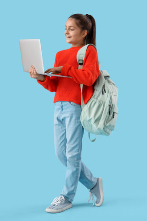 Little schoolgirl with backpack using laptop on blue backgroundの写真素材