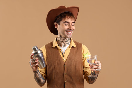 Young male bartender dressed as cowboy with shaker and glass of cocktail on beige backgroundの写真素材