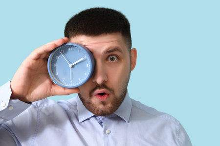 Young shocked businessman with alarm clock showing OK gesture on blue background. Time management conceptの写真素材