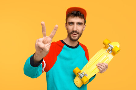Young man with skateboard showing victory gesture on yellow background