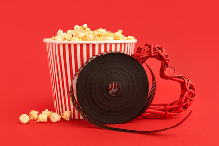 Bucket of popcorn with film reel and decorative heart on red background. Valentine's Day celebrationの写真素材