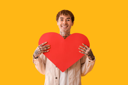 Happy young man with red paper heart on yellow backgroundの写真素材