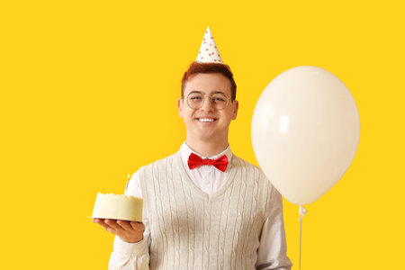 Young man in party hat with air balloon and sweet cake celebrating Birthday on yellow backgroundの写真素材