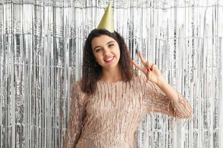 Beautiful young woman in party hat celebrating Birthday and showing victory gesture on tinsel...
