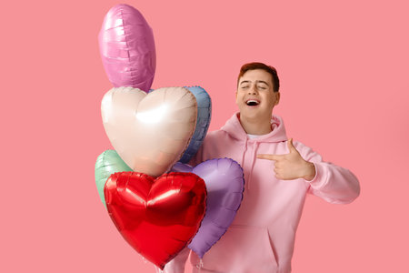 Young man with heart shaped air balloons pointing at something on pink backgroundの写真素材